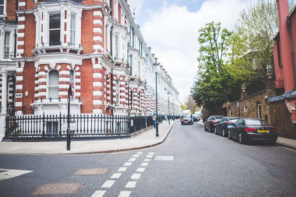 A narrow residential street in an urban area with multi-storey buildings on both sides, featuring classic architectural details, balconies with flower pots, and varied window styles. Several vehicles 