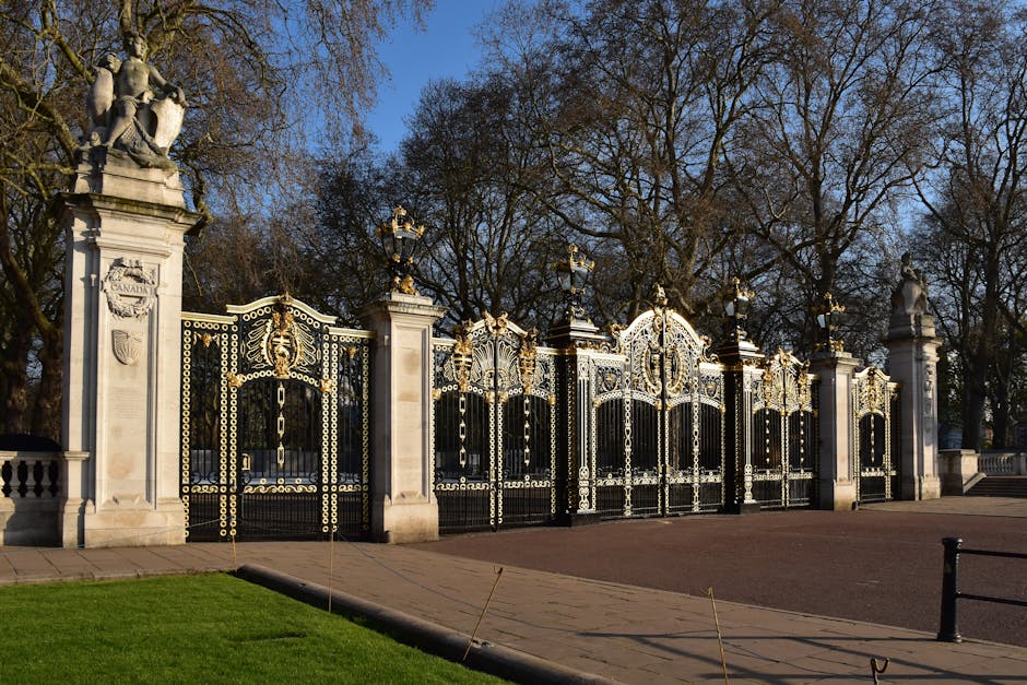 A large ornate gate with intricate black and gold metalwork, featuring decorative scrolls and finials, is flanked by tall stone pillars topped with classical sculptures of seated figures. The gates are partially open, revealing a paved pathway and a row of leafless trees in the background under a clear blue sky. The scene is illuminated by natural sunlight, creating shadows on the pavement and highlighting the detailed craftsmanship of the gate's design. The setting appears to be the entrance to a historic park or estate, with a well-maintained lawn in the foreground. Bayswater Man and Van may utilize such locations for house removals and furniture transport in the Lancaster Gate area near Hyde Park, involving packing, loading, and navigation through similar gates during home relocations.