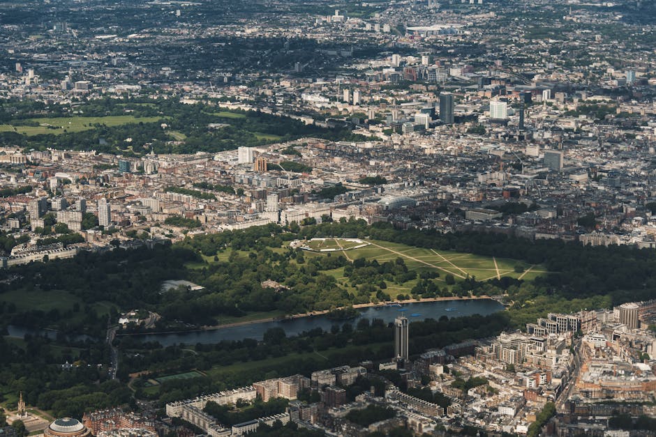 An aerial view of a cityscape showing a large rectangular park with lush green trees, open grassy areas, and pathways, bordered by a river. Surrounding the park are densely packed urban buildings, including high-rise structures, residential blocks, and commercial properties. The image captures the loading process of home relocation, with visible furniture wrapped in protective plastic or fabric being carried by movers, using trolleys and straps to facilitate the transportation. Vehicles belonging to Bayswater Man and Van are seen parked nearby, assisting in furniture transport and packing during the house removal and moving service near Lancaster Gate, close to Hyde Park. The scene depicts a professional moving operation involving careful handling of furniture, loading onto a van, and organizing within the property environment.