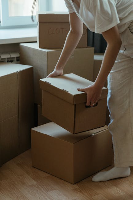A person dressed in a beige top and light-colored trousers is seen inside a room with wooden flooring, preparing to load or move cardboard boxes during a home relocation process. They are carefully handling a box, which is part of a stack of similarly packed boxes, with one marked 'CLOTHES' in red. The boxes are made of plain brown cardboard and are stacked at different heights, some placed directly on the floor, others on top of each other. The scene is well-lit by natural light coming through a window with white frames in the background. This moment appears to be part of a professional removals or packing process, with the individual possibly associated with Bayswater Man and Van, a company providing house removals and furniture transport services. Trolleys, blankets, or packing materials are not visible, and the environment is tidy, emphasizing organized packing and logistics for a house move.