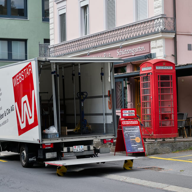 A white removal van with a partially open rear door is parked on the street outside a residential building, with the loading ramp lowered to the pavement. Inside the van, various items such as cardboard boxes, plastic-wrapped objects, and moving equipment like dollies and straps are visible. The van is labeled with the logo of Webstar, indicating a professional relocation service. Adjacent to the van, a traditional red telephone booth stands on the sidewalk near a building with a pink and white facade featuring decorative iron balcony railings and signage in German. The scene suggests preparations for house removals or furniture transport as part of a home relocation process, with the van positioned on the street for efficient loading or unloading of household items, supported by Bayswater Man and Van's professional moving and packing services in the Bayswater area.